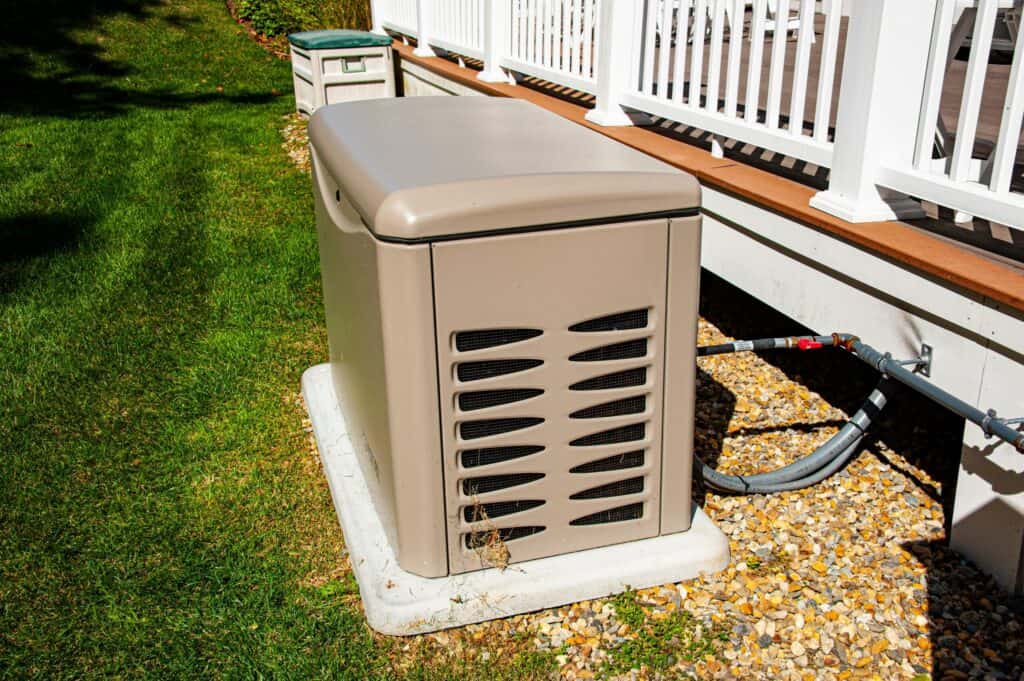 A beige standby generator professionally installed on a concrete pad by the Childers team next to a residential house deck.
