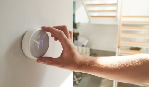 Person's hand adjusting a smart thermostat on a white wall in a home, displaying 77 degrees.